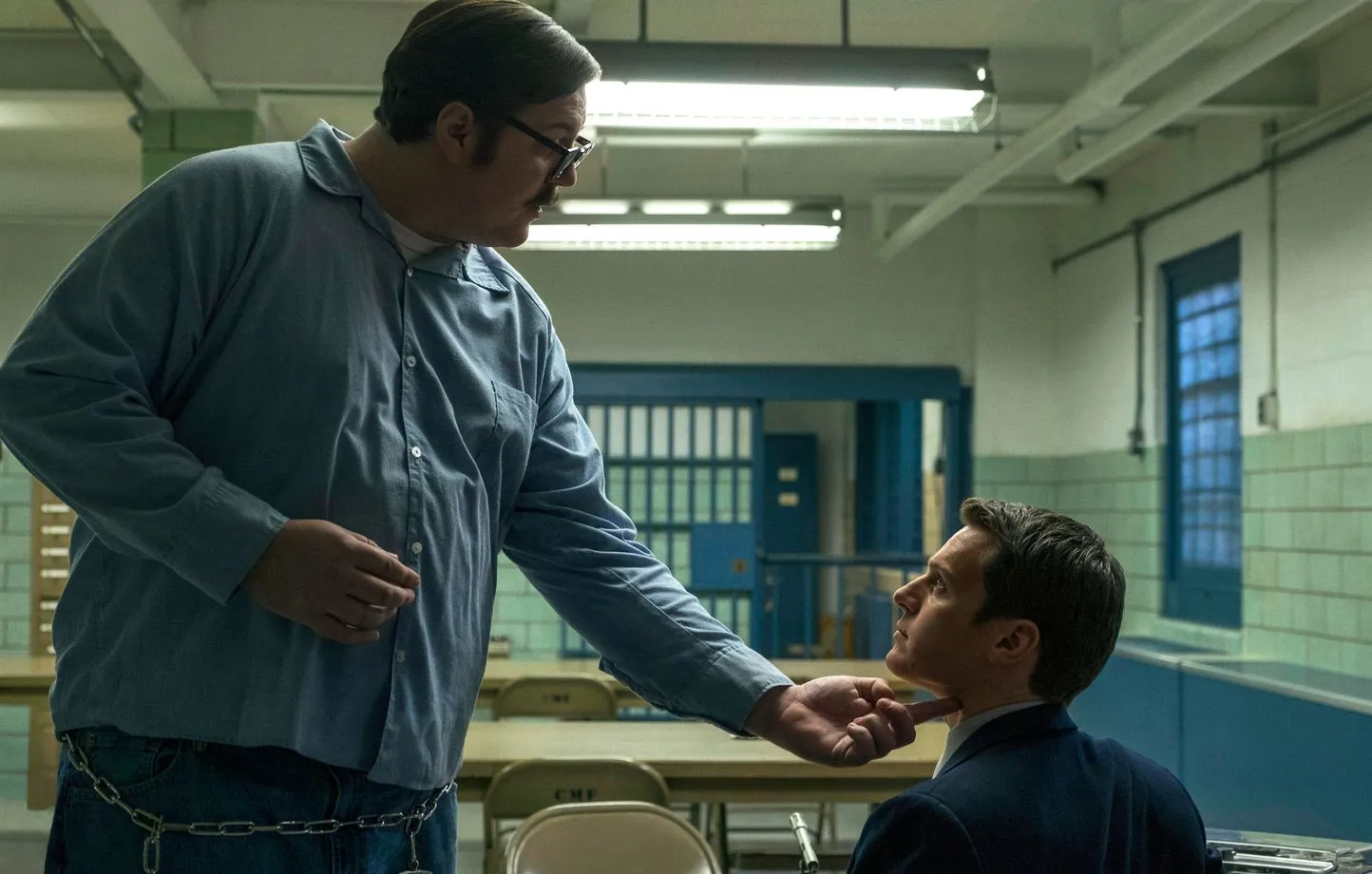 L'acteur Cameron Britton incarnant Edmund Kemper en chemise bleue de prisonnier face à l'agent du FBI dans un parloir. Ils sont tous les deux de profil et le criminel se tient debout face au profiler assis tout en le tenant par le menton.