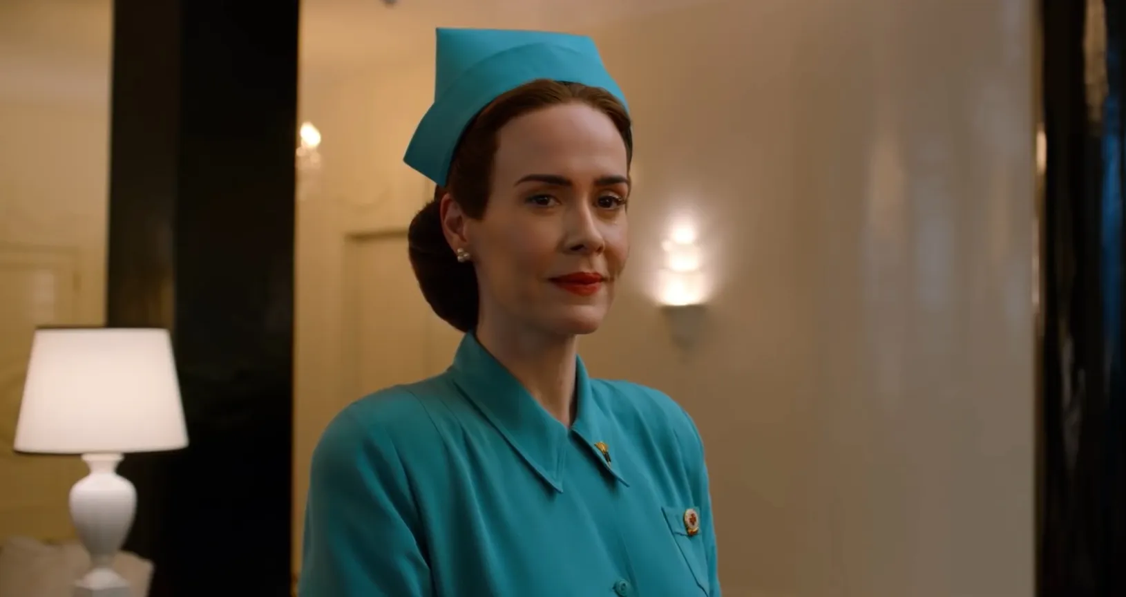 Portrait de l'infirmière Mildred Ratched en uniforme médical vert dans un couloir d'hôpital éclairé. Celle-ci affiche un léger sourire machiavélique et regarde au loin.