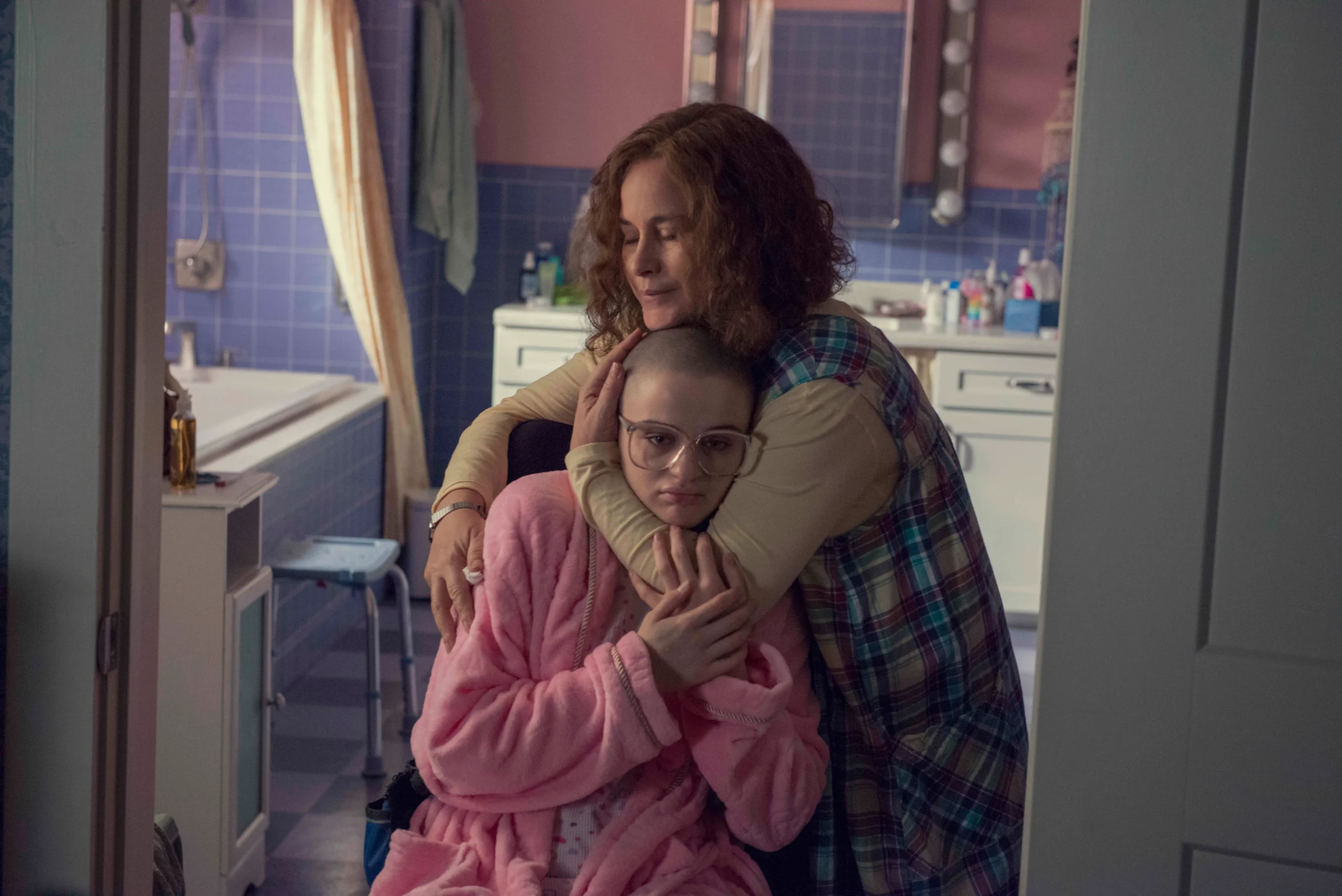 Une mère souriante enlace sa fille en fauteuil roulant dans la salle de bain. Cette dernière a la tête rasée, le visage triste et porte des lunettes et un peignoir rose.