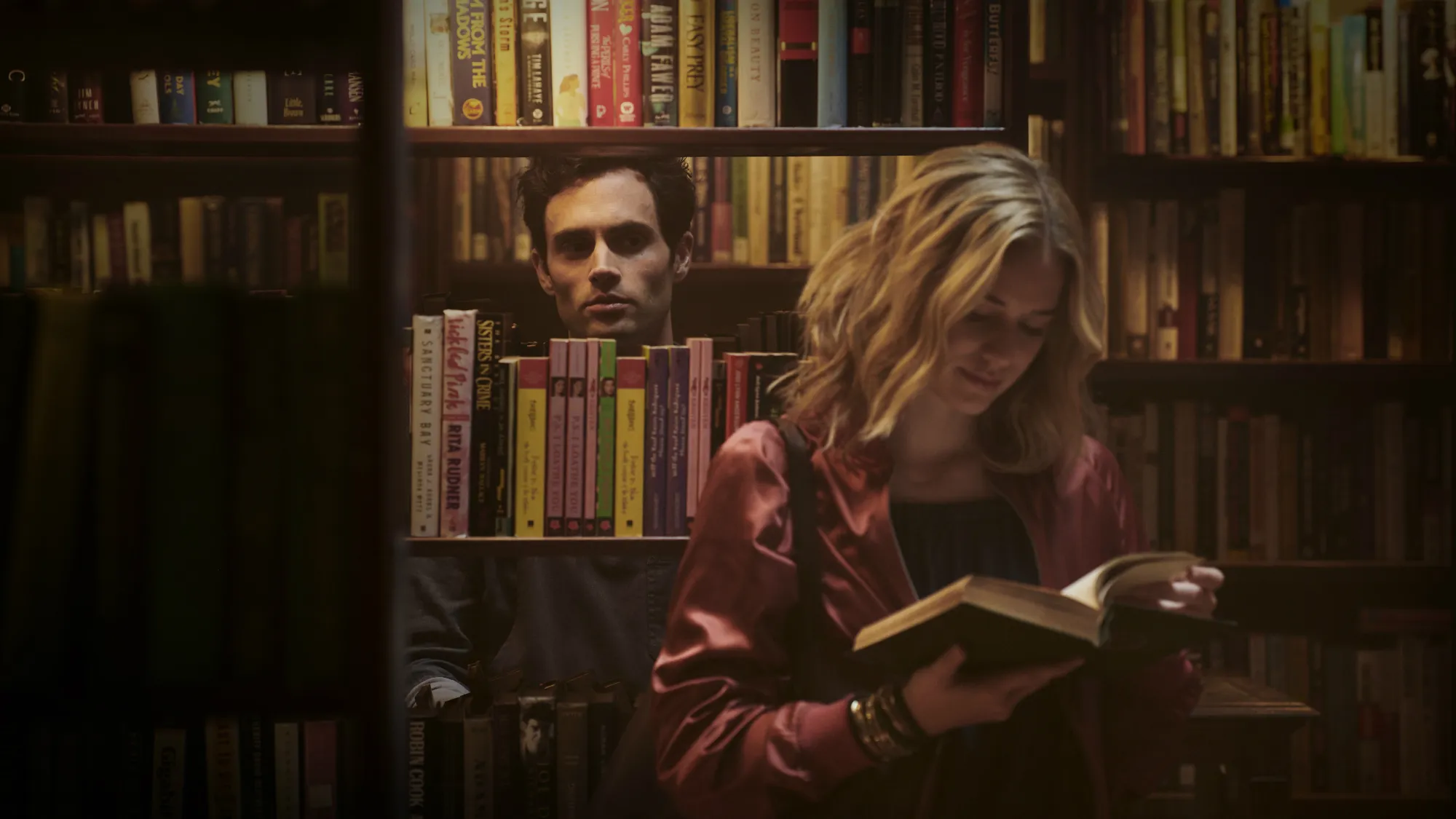 Joe Goldberg observant secrètement Guinevere Beck à travers les étagères d'une bibliothèque sombre pendant qu'elle lit.